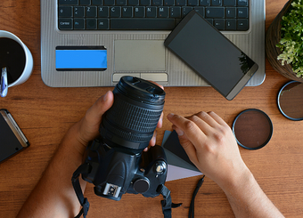 Cropped image of a person holding a camera, laptop and smart, a cup of coffee can also be seen placed on a table