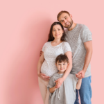 Pregnant mother, father, and son poses for a photo on a pink backgroun
